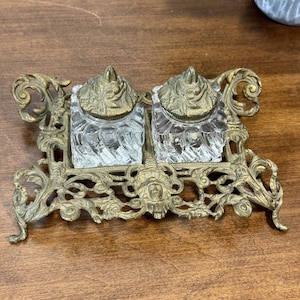 May include: Antique brass inkwell set with two clear glass ink pots. The ink pots have ornate brass lids and sit in a decorative brass tray with a face design. The inkwell is on a wooden surface.