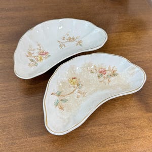 Puede incluir: Dos platos de color blanco roto con forma de riñón con diseños florales en rosa, amarillo y verde. Los platos tienen un borde dorado y están colocados sobre una superficie de madera. El estampado floral es delicado y de inspiración vintage.