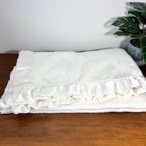 May include: A soft, off-white fleece blanket featuring a smooth, shiny satin trim along the edges. The blanket is folded neatly, showcasing the textured fabric of the main body contrasted against the lustrous, ruffled satin border detail.