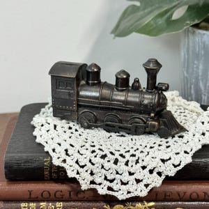 May include: A miniature bronze-colored metal steam locomotive train figurine featuring a detailed boiler, a tall smokestack, a cab, and visible side wheels. The train has a weathered, dark metallic finish with subtle highlights on the raised edges.