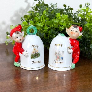 May include: Two white ceramic bell figurines featuring painted elves in red outfits. The left bell shows a candle wreath and the text Merry Christmas. The right bell displays hanging ornaments and the text Merry Christmas in gold lettering.