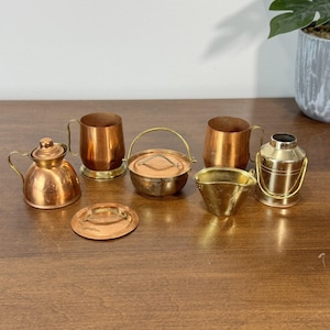 May include: A collection of miniature copper and brass kitchenware items, including a teapot, mugs, a small pot with a handle, a bucket, and a small bowl. The items are arranged on a wooden surface.