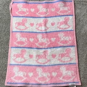 Könnte beinhalten: Rosa-weißes Babydecke mit einem Schaukelpferdmuster. Die Decke hat abwechselnde Streifen in Rosa und Weiß mit blauen Akzenten. Die Schaukelpferde und Herzen sind weiß. Die Decke hat einen rosa Rand und eine Schlaufe.
