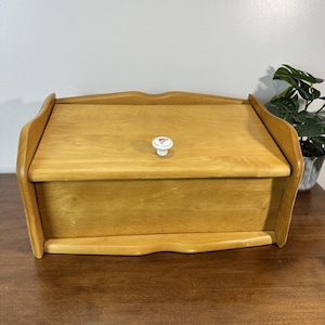 May include: A rectangular wooden bread bin with a light brown finish and a curved top edge. The front features a drop-down door with a small, round white ceramic knob centred on the front panel, decorated with a tiny pink floral pattern.
