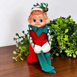 May include: A vintage-style elf doll with a pale face, blue eyes, and rosy cheeks. The doll wears a red felt body, a green collar, and a white and green striped hat topped with a small metal bell. The elf has white felt mittens and long green felt sleeves.