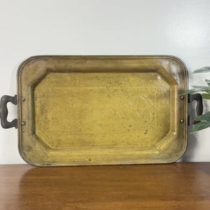 Puede incluir: Una bandeja rectangular de latón con un patrón floral decorativo y asas de madera oscura. La bandeja tiene un tono dorado y está colocada sobre una superficie de madera. Las asas están sujetas con pequeños remaches de latón.