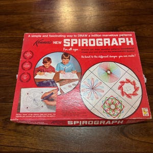 May include: A red and white box for the Kenner's New Spirograph drawing toy. The box shows two children drawing with the Spirograph and examples of the designs that can be created. The text on the box reads "A simple and fascinating way to DRAW a million marvelous patterns Kenner's NEW SPIROGRAPH For all ages... Anyone can draw beautiful patterns immediately! Create your own and use the SPIROGRAPH Booklet No limit to the different designs you can make!"