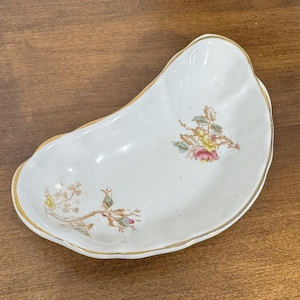 May include: A white, kidney-shaped ceramic dish with a gold rim and floral accents. The dish features delicate illustrations of pink, yellow, and green flowers and leaves. The dish is likely vintage or antique.