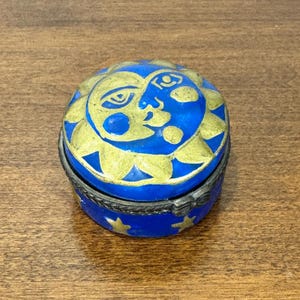 May include: A small, round blue trinket box with a hinged lid. The lid features a gold sun face design. The box's base is decorated with gold stars. The box has a silver-colored metal band around the middle.