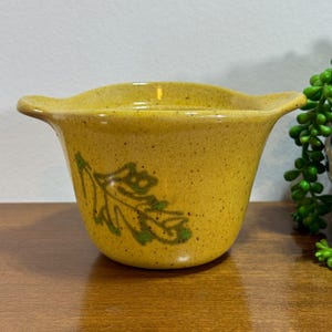 以下が含まれることがあります： 緑の葉のデザインが施された黄色の陶器製のプランター。プランターは斑点のある釉薬と丸みを帯びた形をしています。