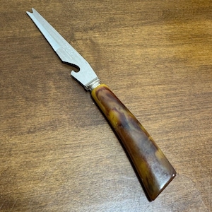 May include: A vintage multi-tool with a stainless steel blade and bottle opener. The handle is a marbled brown and yellow color. The tool is designed for cutting and opening bottles.