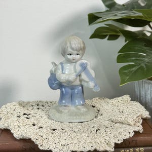 Puede incluir: Figura de porcelana de un niño tocando el violín. La figura lleva un peto azul y una camisa blanca. El violín es azul claro y blanco. La figura está sobre una base pequeña y redonda.