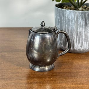 Puede incluir: Una pequeña tetera antigua de color plateado con tapa y asa. La tetera tiene un cuerpo redondeado y se asienta sobre una pequeña base. El asa es curva y la tapa tiene un pequeño pomo en la parte superior. La tetera está colocada sobre una superficie de madera.