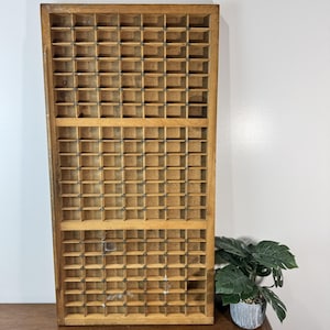 May include: A vintage wooden printer's tray with numerous small, square compartments. The tray is made of light-colored wood and has a rectangular shape. It is designed to hold individual letters or other small items. A potted plant is visible in the background.