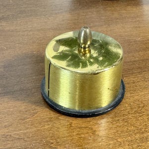 Pode incluir: Um recipiente cilíndrico dourado com um pequeno topo arredondado e uma base preta. O recipiente tem um pequeno botão dourado no topo. O objeto está sobre uma superfície de madeira.