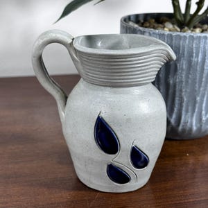 May include: A light gray ceramic pitcher with a curved handle and a ribbed neck. The body of the pitcher features three dark blue, teardrop-shaped indentations arranged in a cluster on the front surface, providing a distinct contrast.