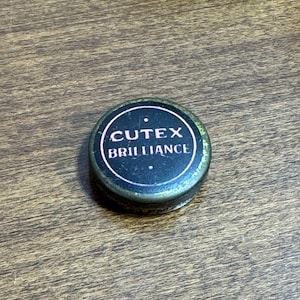 May include: A small, round, metal container with a dark blue top. The top features the words "CUTEX BRILLIANCE" in white, within a white circular border. The container sits on a wooden surface.