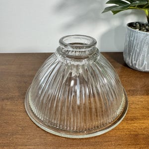 May include: Clear glass lampshade with a ribbed, bell-shaped design. The lampshade has a wide base and a narrow top, with a slightly textured rim. The glass is transparent, allowing light to pass through. The lampshade is sitting on a wooden surface.