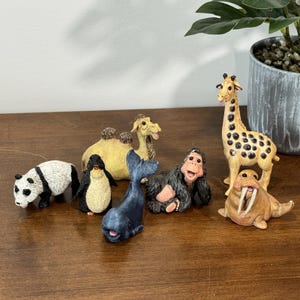 May include: A collection of miniature animal figurines, including a panda, penguin, whale, gorilla, camel, giraffe, and walrus. The figurines are painted in realistic colors and are displayed on a wooden surface.