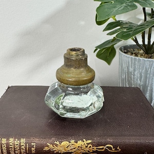 Op de afbeelding: Een inktpot van helder glas met een gefacetteerde, meerzijdige basis en een verweerde, doffe koperen kraag aan de bovenkant. Het glas is transparant met zichtbare lichtreflecties en het metalen onderdeel vertoont tekenen van ouderdom en oxidatie.