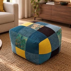 Op de afbeelding: Een vierkante vloerpoef met een patchworkontwerp met diverse stoftexturen en kleuren, waaronder mosterdgeel, diep petrol, donker marineblauw en een gespikkeld groen en goud patroon, uitgevoerd in een gestructureerde, lage kubusvorm.