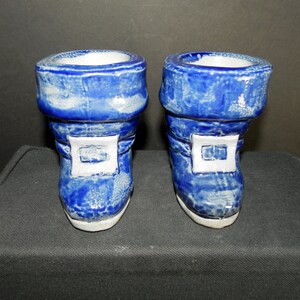 May include: A pair of ceramic boot-shaped containers featuring a glossy blue glaze with white mottled accents. Each boot has a prominent white square buckle detail on the front, a wide cuff at the top, and a smooth white base, creating a uniform set.
