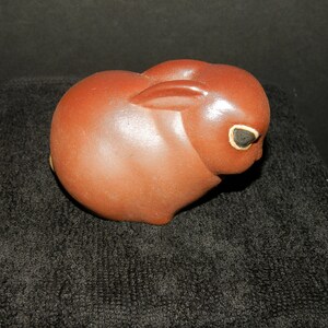 May include: A smooth, reddish-brown ceramic figurine shaped like a crouching rabbit. The rabbit has a rounded body, ears laid flat against its back, and dark, almond-shaped eyes outlined in a light, contrasting color. The surface has a subtle sheen.