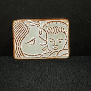 May include: A rectangular ceramic plaque featuring a light cream-colored glaze with brown line work depicting a profile of a person kissing the forehead of a child. The edges of the plaque are finished in a matching brown tone.