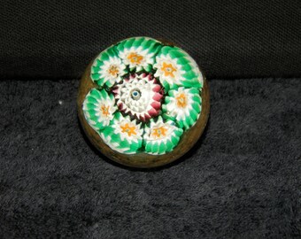 Puede incluir: Un pisapapeles de vidrio esférico con un diseño millefiori que presenta múltiples patrones florales en verde, blanco y naranja dispuestos en círculo alrededor de un grupo central de elementos de vidrio blanco y rojo oscuro.