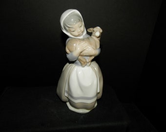 May include: A glossy porcelain figurine of a young person wearing a white head covering and a long dress with a white apron, holding a small light brown lamb in their arms. The figurine features soft grey, white, and tan colour tones throughout.