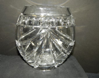 Puede incluir: Un jarrón esférico de cristal transparente con un borde ancho y liso, y un cuerpo decorado con profundas ranuras de vidrio tallado vertical que irradian desde la base, creando un patrón en forma de abanico alrededor del centro.