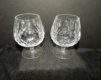 Puede incluir: Dos copas de brandy de cristal transparente con un patrón de corte de diamante profundo en la mitad inferior de los cuencos. Cada copa tiene un tallo corto y grueso y una base circular con un diseño de estrella radiante grabado.