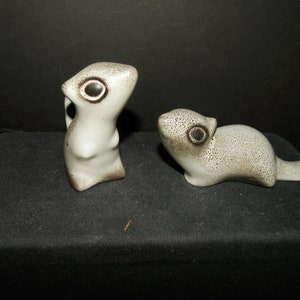 Puede incluir: Dos figuras de animales de cerámica con un esmalte suave de color blanco roto y detalles moteados en marrón oscuro. Una figura está en posición vertical, mientras que la otra está en posición horizontal. Ambas tienen ojos grandes, oscuros y circulares.
