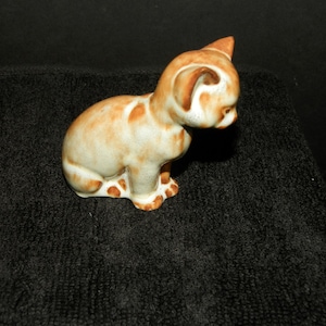 May include: A ceramic cat figurine in a seated position, featuring a cream-colored base with mottled orange and brown patches across its body, head, and ears. The cat is depicted looking downward with its head tilted toward the ground.