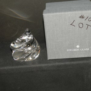 May include: A clear, swirl-shaped glass sculpture with a smooth, reflective surface. Beside it sits a gray, textured, square box with the text #109 LOTA written in black marker and the Steuben Glass logo printed in white on the front face.