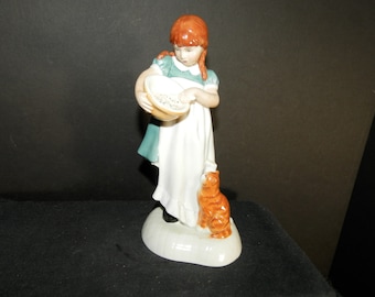 May include: A porcelain figurine of a young person with reddish-brown hair in braids, wearing a teal dress with a white apron, holding a small bowl while looking down at an orange tabby cat sitting on a white oval base at their feet.
