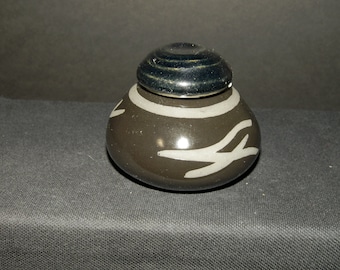 Può includere: Un piccolo vasetto in ceramica arrotondato con una finitura marrone scuro lucida. Il corpo presenta un'ampia fascia orizzontale grigio chiaro vicino alla parte superiore e segni astratti a forma di ramo di colore grigio chiaro.