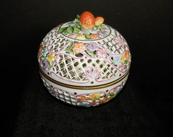 May include: A round porcelain lidded bowl featuring an intricate white lattice design with gold accents. The surface is decorated with hand-painted colorful flowers and green leaves. The lid is topped with a sculpted orange fruit finial.