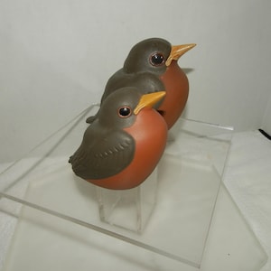 May include: Two robin bird figurines with dark brown wings and backs, orange-red chests, and yellow beaks. The birds have dark, glossy eyes and are positioned one behind the other, featuring smooth, matte-textured surfaces on their bodies.
