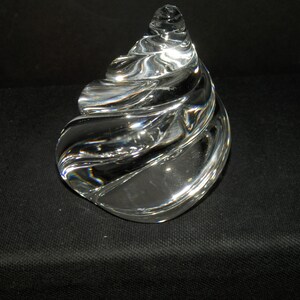 May include: A clear glass paperweight shaped like a spiral seashell. The object features a smooth, polished surface with curved, translucent ridges that catch the light, creating a reflective and transparent appearance throughout the entire form.
