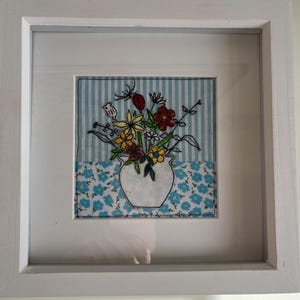 May include: A framed embroidery of a white vase filled with colourful flowers on a blue and white striped background.