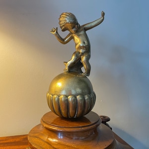 May include: A gold-toned metal sculpture of a cherub standing on a sphere with a decorative base. The cherub is depicted with wings and a playful expression.