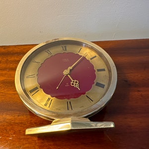 May include: A gold-toned Oris 7 Jewels alarm clock with a red face and Roman numeral hour markers. The clock has a day display and is made in Switzerland.