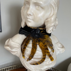 May include: A white marble bust of a young girl wearing a black velvet choker with a black lace bow and a brown and gold feather accent.
