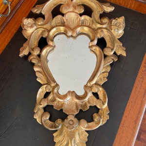 Puede incluir: Un espejo de madera pintado en oro con un diseño ornamentado e intrincado. El espejo tiene forma de corazón y tiene un borde biselado.