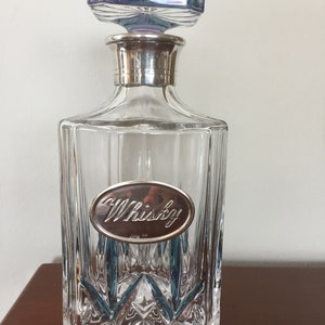 Puede incluir: Una decantador de vidrio transparente con un tapón de plata y una etiqueta de plata que dice "Whisky". La decantador tiene un patrón texturizado en forma de diamante.