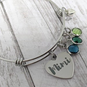 May include: A silver expandable bracelet with a heart charm engraved with the name "Mimi". The bracelet has four dangling charms with green and blue gemstones.