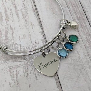 May include: A silver expandable bangle bracelet with a heart charm engraved with the name "Hanna". The bracelet has three blue and green crystal charms.