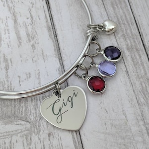 May include: A silver expandable bracelet with a heart charm engraved with the word "Gigi". The bracelet also features three dangling charms: a small heart, a purple crystal, and a red crystal.