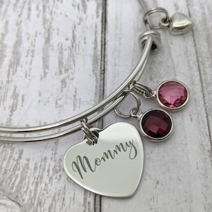 May include: A silver expandable bangle bracelet with a heart charm engraved with the word "Mommy". The bracelet also features two dangling charms: a pink crystal and a burgundy crystal.